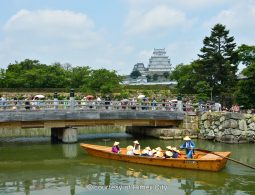 Himeji Castle seen from across the moat, with a sightseeing boat carrying people in straw hats passing beneath a pedestrian bridge.