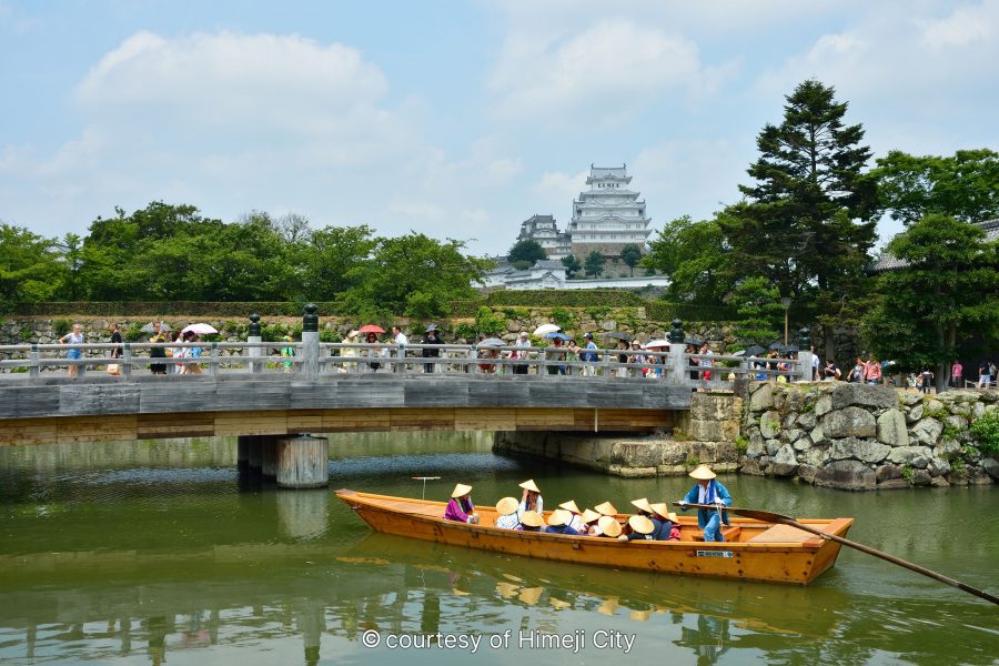 Himeji Castle seen from across the moat, with a sightseeing boat carrying people in straw hats passing beneath a pedestrian bridge.