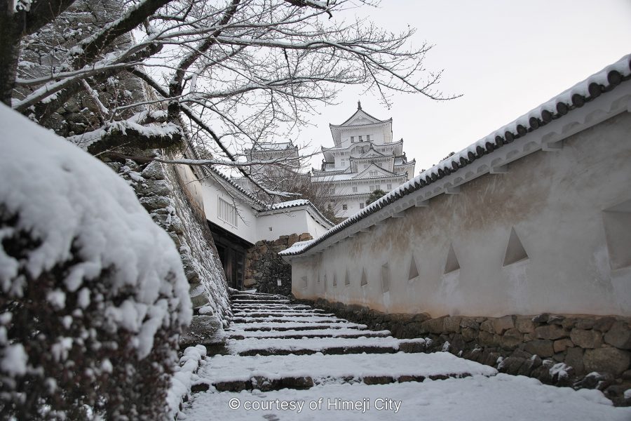 Snow-covered stone steps and white walls leading up toward Himeji Castle on a cloudy winter day.