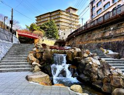 A small, tiered waterfall flowing over rocks, flanked by stone stairs and a red bridge in the Arima Onsen town center, with modern buildings in the background.