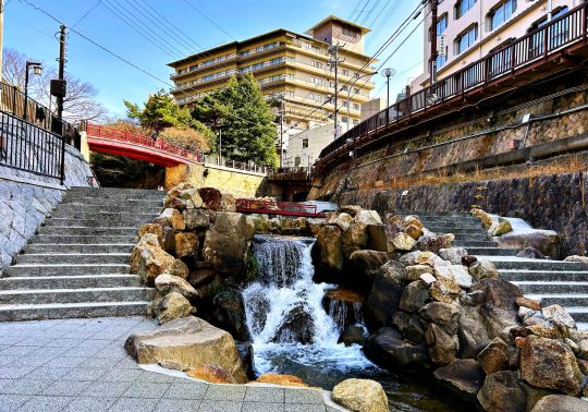 A small, tiered waterfall flowing over rocks, flanked by stone stairs and a red bridge in the Arima Onsen town center, with modern buildings in the background.