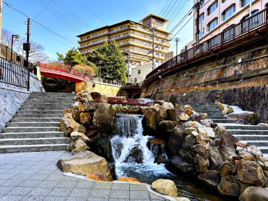 A small, tiered waterfall flowing over rocks, flanked by stone stairs and a red bridge in the Arima Onsen town center, with modern buildings in the background.