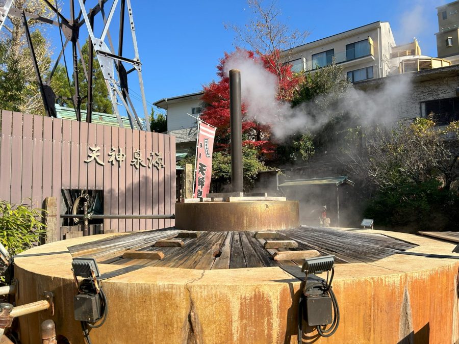 A large, steaming hot spring source display (Tensin Sen Gen) in Arima Onsen, with a tall pipe emitting steam and red maple leaves in the background.