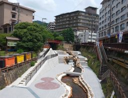 A wide, modern river channel running through the Arima Onsen town center, flanked by stone walls, stairs, and modern hotels.