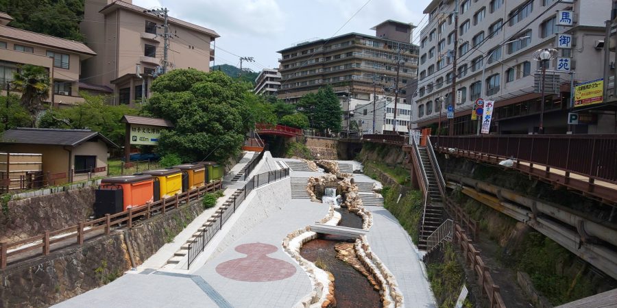 A wide, modern river channel running through the Arima Onsen town center, flanked by stone walls, stairs, and modern hotels.
