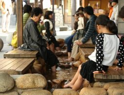 People sitting on wooden benches dipping their feet in a warm public foot bath (ashiyu) in Arima Onsen, surrounded by smooth river stones.