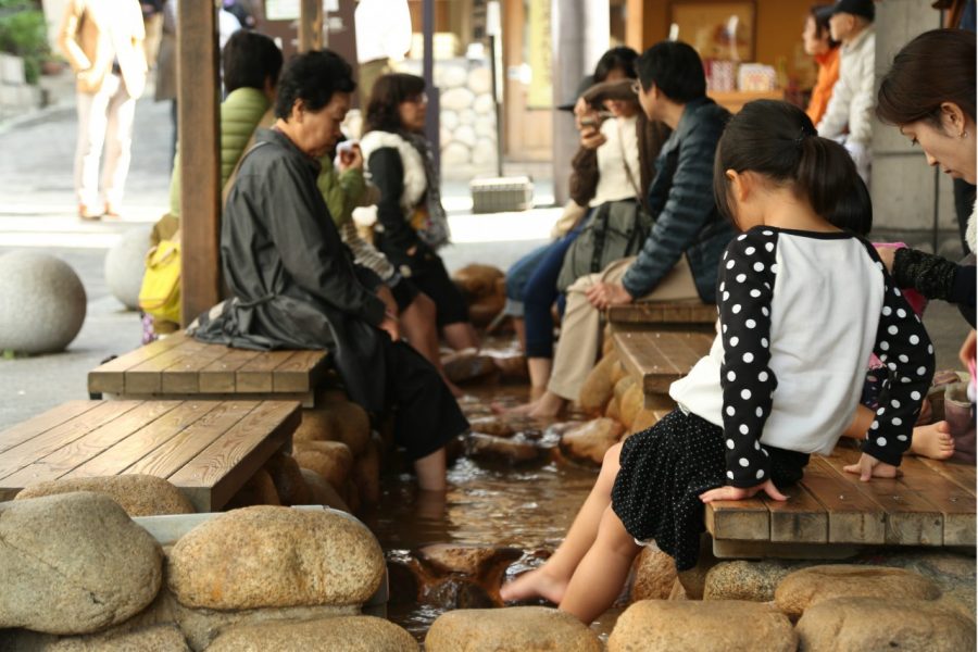 People sitting on wooden benches dipping their feet in a warm public foot bath (ashiyu) in Arima Onsen, surrounded by smooth river stones.