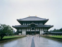 The large, imposing wooden structure of the Todai-ji Great Buddha Hall (Daibutsuden) viewed from the pathway, with a dark tiled roof and golden horns.
