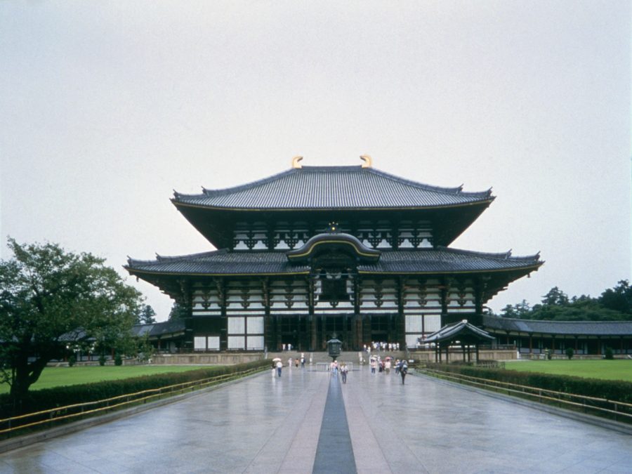 The large, imposing wooden structure of the Todai-ji Great Buddha Hall (Daibutsuden) viewed from the pathway, with a dark tiled roof and golden horns.