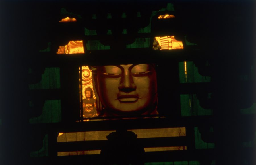 A close-up of the face of the Great Buddha statue (Daibutsu) seen dimly through dark wooden lattice work, illuminated by warm, golden light.