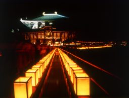 A dramatic night view of the illuminated Todai-ji Daibutsuden, with a long pathway lined by glowing square paper lanterns.