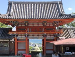 The two-story Romon Gate of Kishu Toshogu, showing its dark tiled roof, red framework, and a view of the sea through the gate opening.