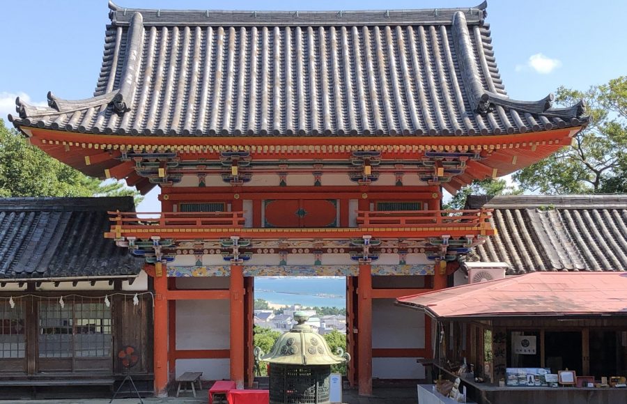 The two-story Romon Gate of Kishu Toshogu, showing its dark tiled roof, red framework, and a view of the sea through the gate opening.