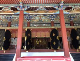 Interior of the Kishu Toshogu Shrine Haiden (Worship Hall), showing detailed, colorful carvings on the ceiling beams and four large black panels with gold accents.
