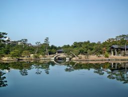 A scenic view across the pond of Yosuien Garden, showing an arched bridge and pavilion reflected in the calm water.