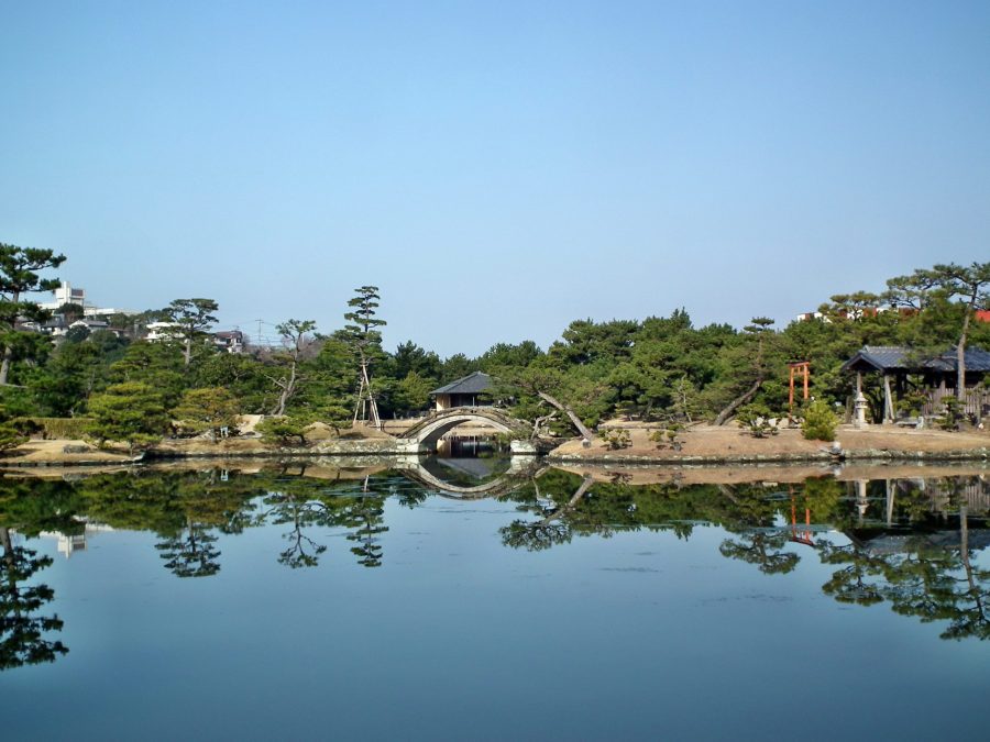 A scenic view across the pond of Yosuien Garden, showing an arched bridge and pavilion reflected in the calm water.