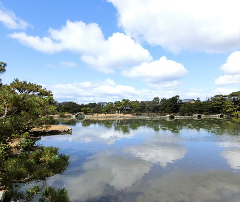 The large central pond (tidal pond) of Yosuien Garden, with arched stone bridges, reflecting the sky and surrounding trees.