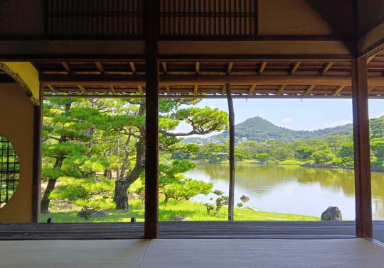 View of the Yosuien Garden pond and manicured pine trees from inside a traditional wooden building's veranda.