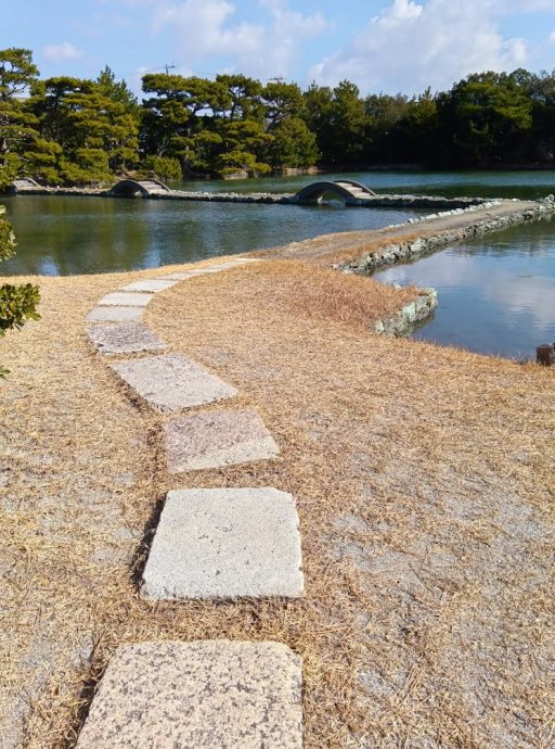 A stone path leading toward the pond in Yosuien Garden, with the arched bridges visible in the distance.