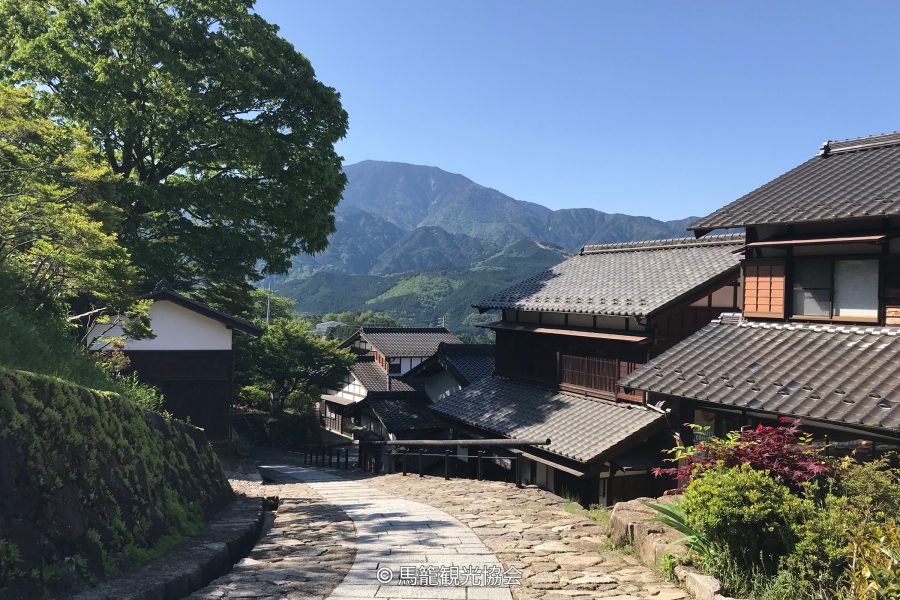 The stone-paved, sloping main street of the Magome-juku post town, lined with traditional wooden buildings and green trees, with mountains in the background.