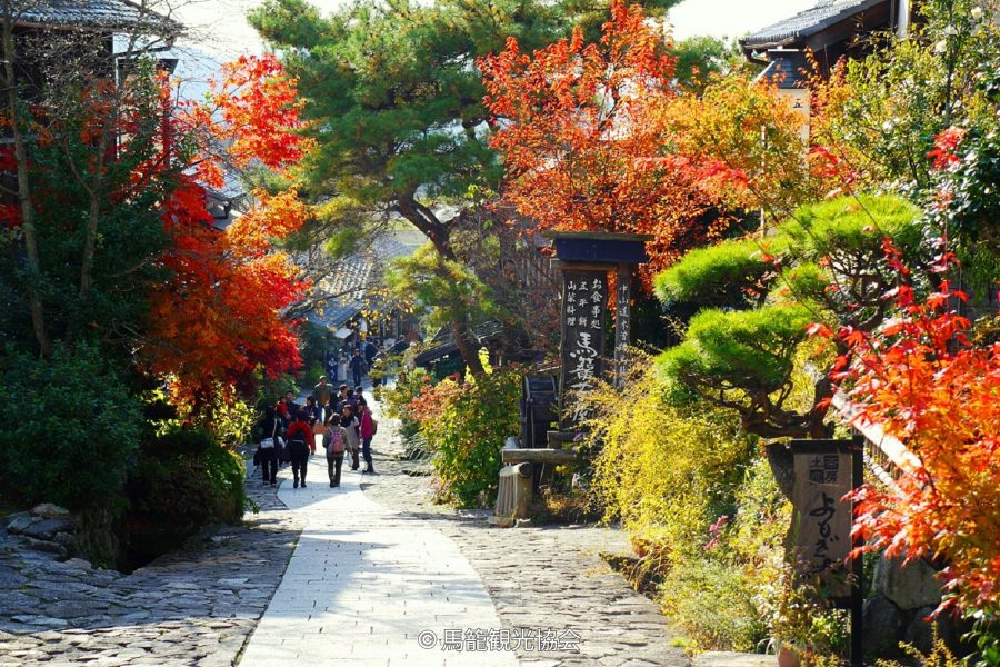 The sloping stone-paved street of Magome-juku, surrounded by vibrant red and orange autumn foliage with visitors walking uphill.