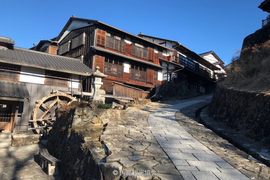 Traditional dark wood houses lining the sloping cobblestone street of Magome-juku, with a wooden water wheel visible on the left.