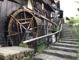 A large wooden water wheel and a flight of stone stairs ascending the hillside next to a traditional building in Magome-juku.