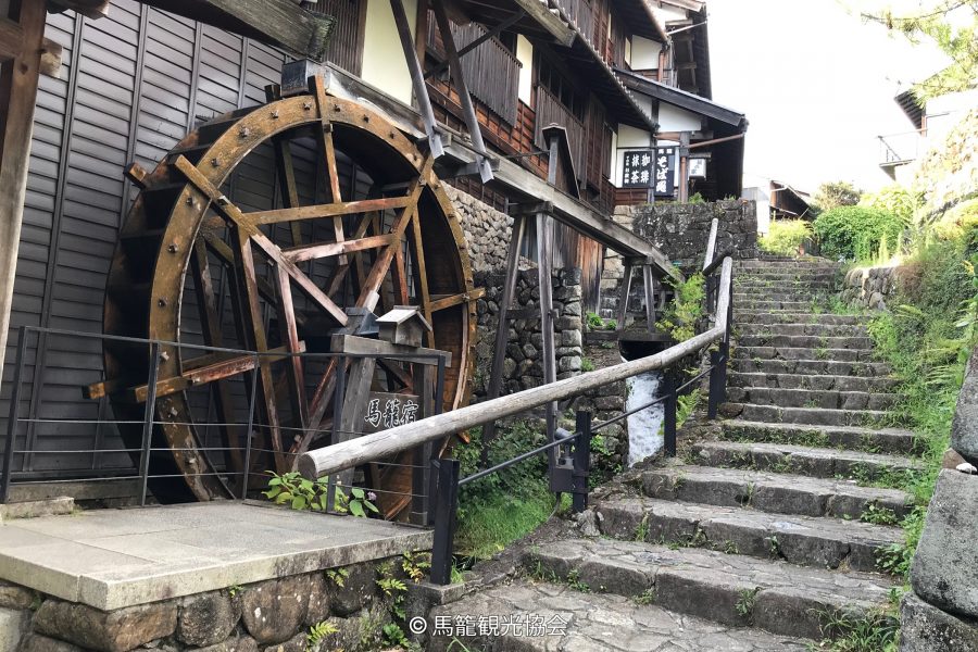 A large wooden water wheel and a flight of stone stairs ascending the hillside next to a traditional building in Magome-juku.