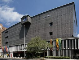 The modern exterior of the National Bunraku Theatre in Osaka, featuring a dark tiled facade and vertical white slats on the lower level, with multi-colored banners hanging in front.