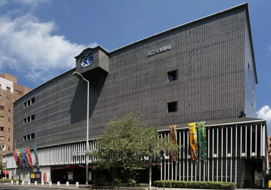 The modern exterior of the National Bunraku Theatre in Osaka, featuring a dark tiled facade and vertical white slats on the lower level, with multi-colored banners hanging in front.