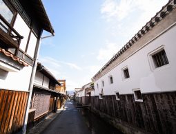 A narrow street lined with white-walled storehouses and wooden merchant houses, with a small canal running alongside it.
