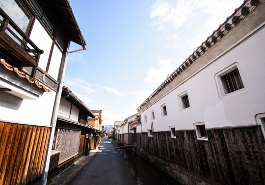 A narrow street lined with white-walled storehouses and wooden merchant houses, with a small canal running alongside it.