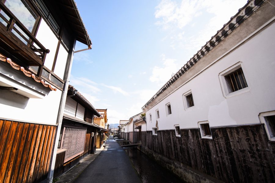 A narrow street lined with white-walled storehouses and wooden merchant houses, with a small canal running alongside it.
