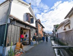 A close-up of the Kurayoshi district with its white-walled buildings, showing a cafe/shop next to a small canal, with two people walking away.