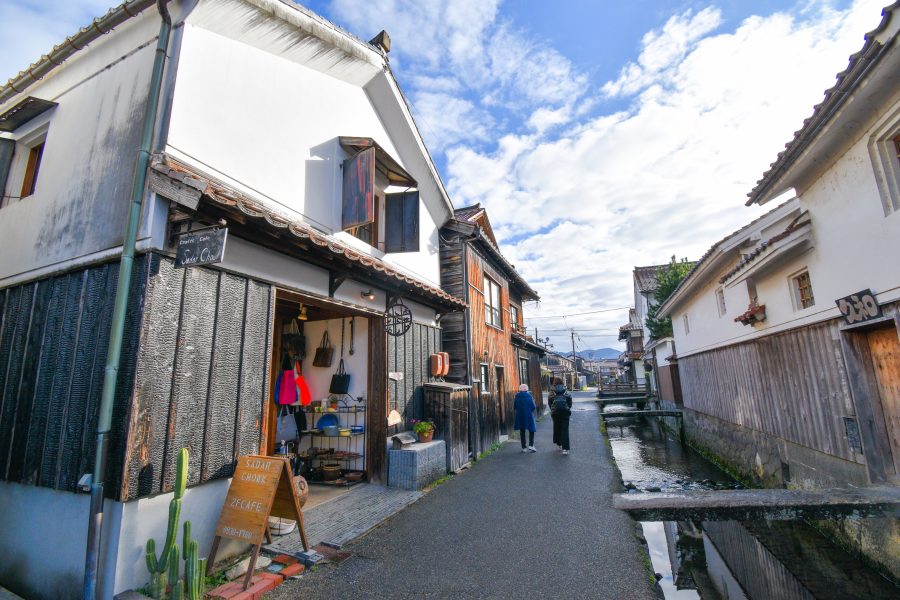 A close-up of the Kurayoshi district with its white-walled buildings, showing a cafe/shop next to a small canal, with two people walking away.