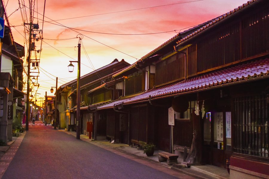 The Kurayoshi white-walled district streetscape at dusk, illuminated by warm lights under a dramatically orange-red sky.