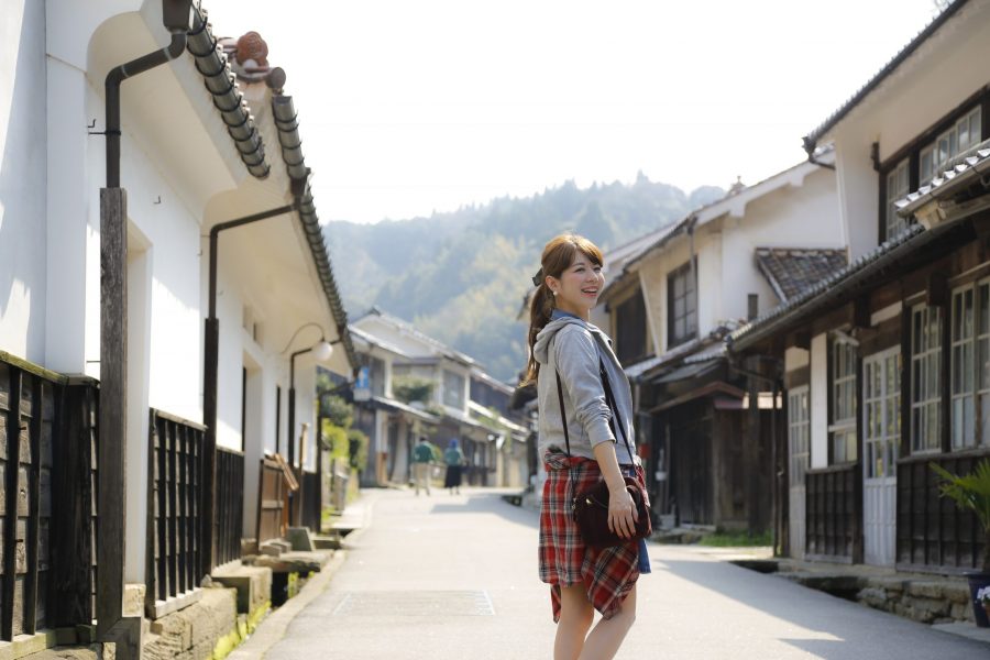 A woman standing on the preserved Omori townscape street of the Iwami Ginzan Silver Mine area, with white-walled houses and forested hills in the background.