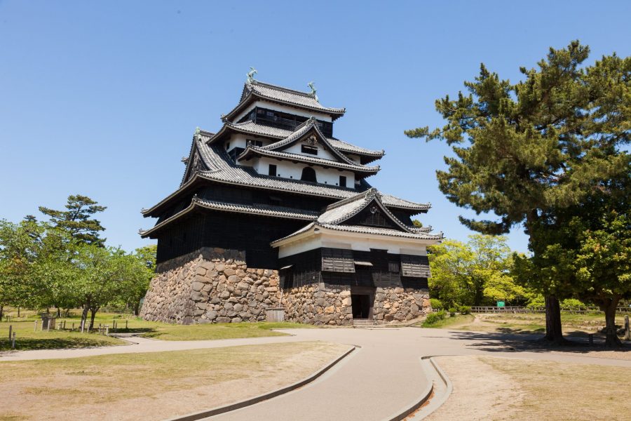 The black-walled Matsue Castle keep and stone base, standing on a grassy hill under a clear blue sky.