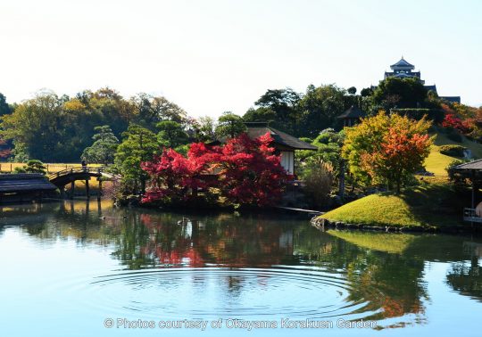 Autumn view of Okayama Korakuen Garden's central pond, reflecting vibrant red maple leaves and a small teahouse, with Okayama Castle visible on the hill in the distance.