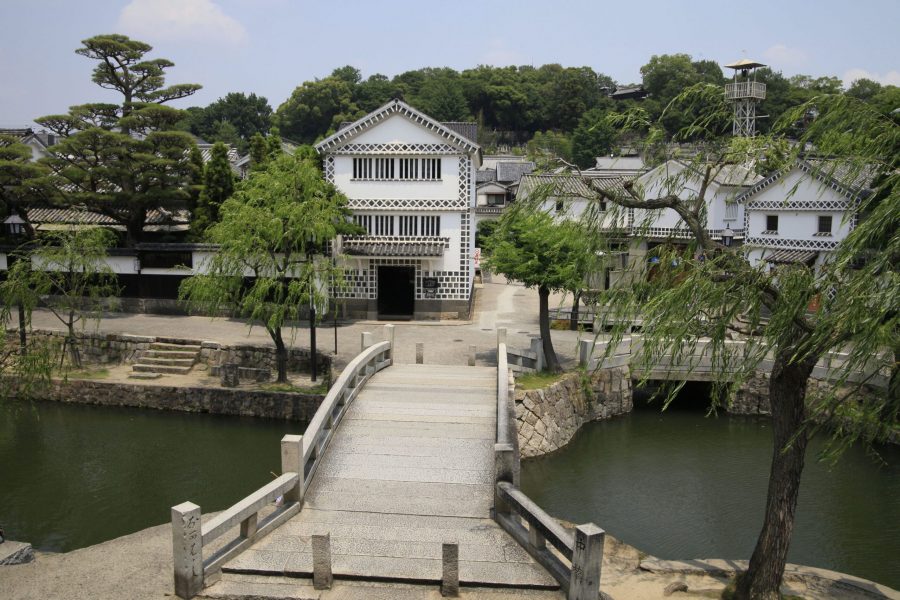 An elevated view over a stone bridge of the white-walled Kurashiki Bikan Historical Area, with a two-story building ahead.