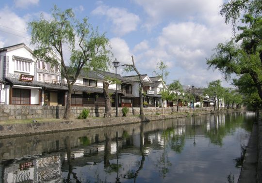 A wide shot of the Kurashiki Canal (Kurashiki River) reflecting the white-walled historical houses and weeping willow trees under a partially cloudy sky.