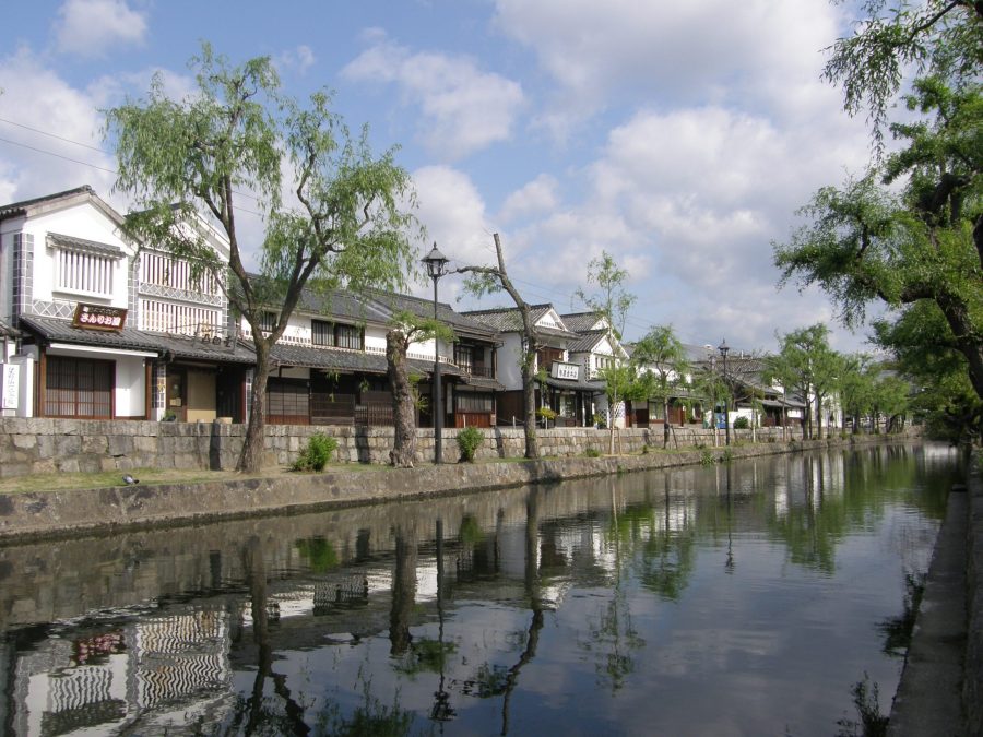 A wide shot of the Kurashiki Canal (Kurashiki River) reflecting the white-walled historical houses and weeping willow trees under a partially cloudy sky.