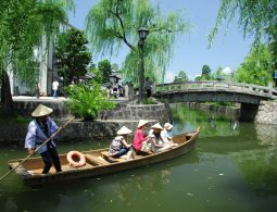 A traditional flat-bottom boat carrying passengers with straw hats on the Kurashiki River, passing under a stone bridge.