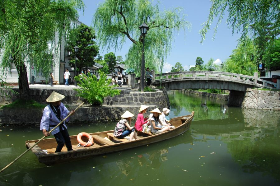 A traditional flat-bottom boat carrying passengers with straw hats on the Kurashiki River, passing under a stone bridge.