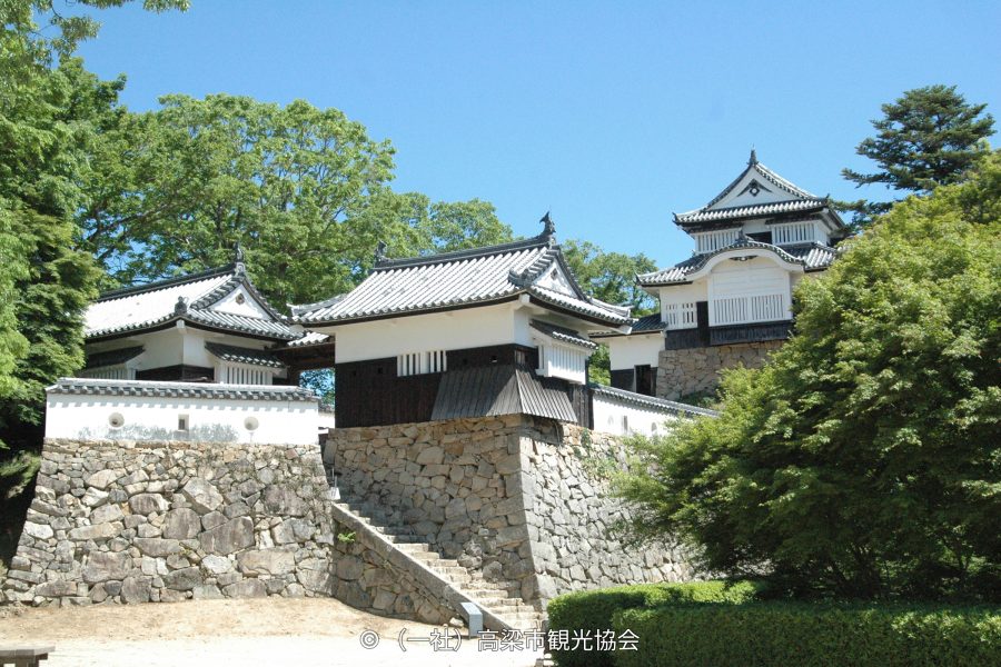 The main castle buildings and connecting structures (Yagura) of Bitchu Matsuyama Castle on a stone wall, surrounded by lush green trees.