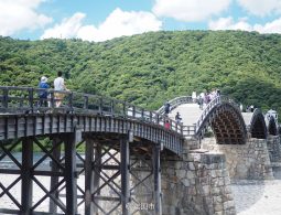 People walking across the historic wooden Kintai Bridge with its five distinct arches and stone piers, backed by a green, wooded hill.