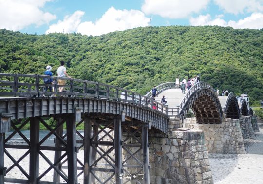 People walking across the historic wooden Kintai Bridge with its five distinct arches and stone piers, backed by a green, wooded hill.