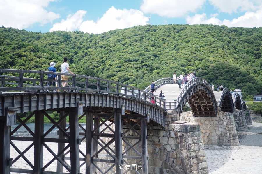 People walking across the historic wooden Kintai Bridge with its five distinct arches and stone piers, backed by a green, wooded hill.