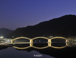 The illuminated Kintai Bridge at dusk, with its five arches and their reflections creating a series of perfect circles on the calm water.
