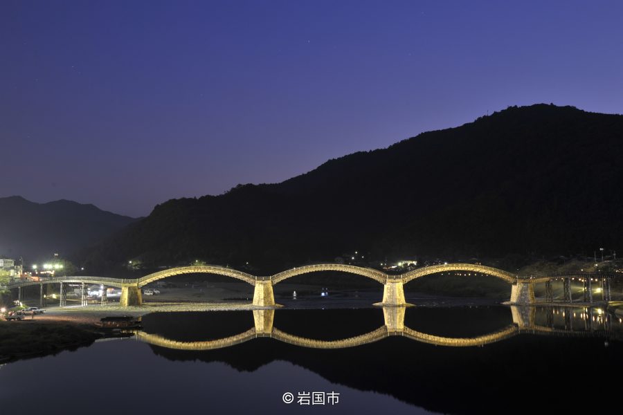 The illuminated Kintai Bridge at dusk, with its five arches and their reflections creating a series of perfect circles on the calm water.
